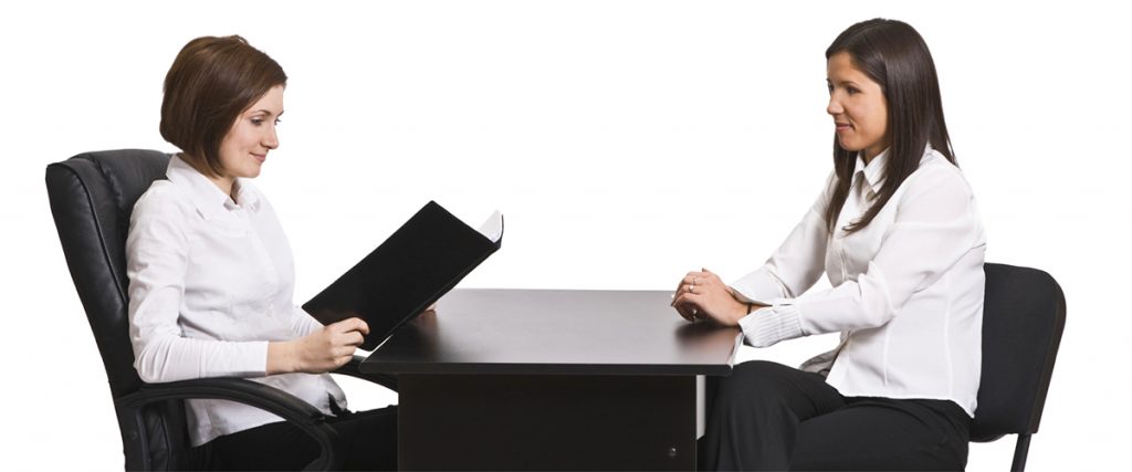 two women sitting a table with one interviewing the other, mid thirties in age