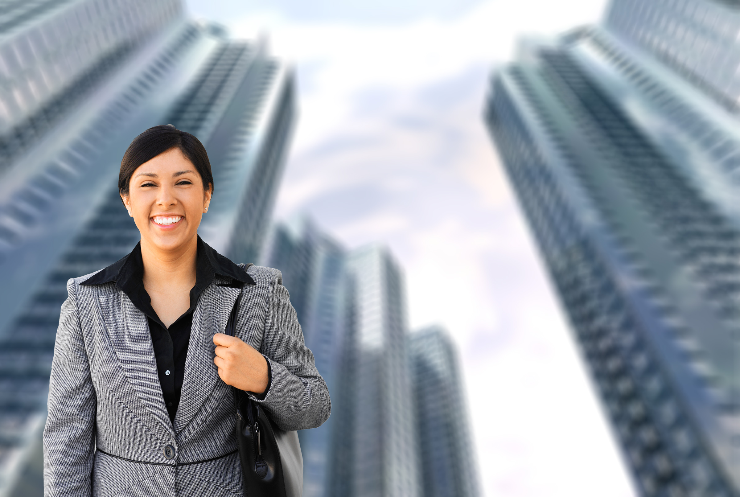 success story - confident female executive, mid forties, arms down, gray suit, black blouse, smiling, superimposed on blurred background of tall skyscrapers